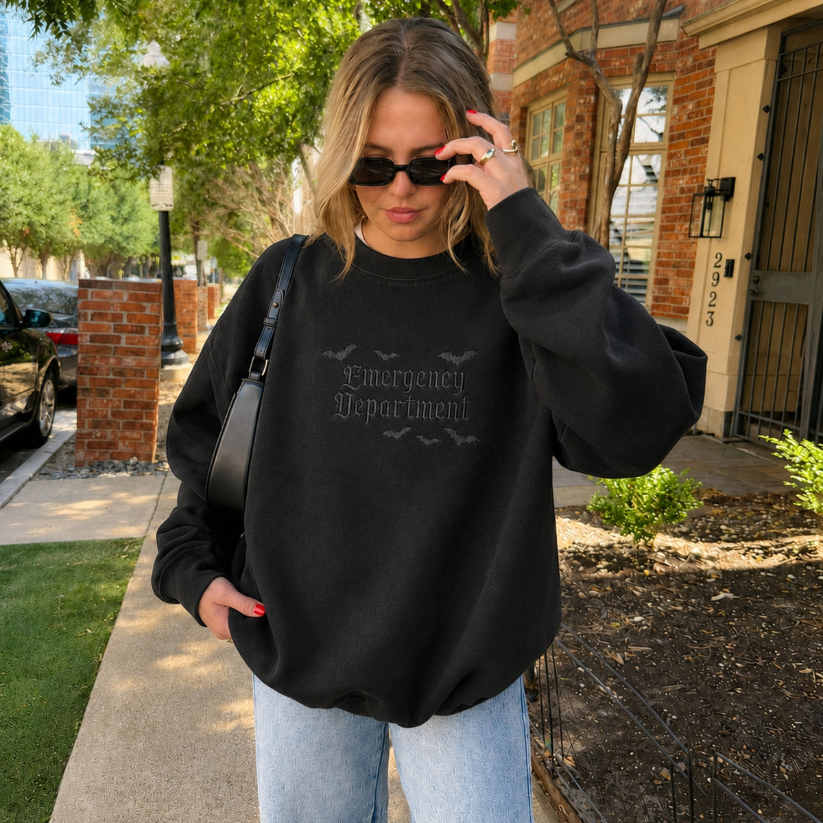A woman wearing a black crewneck sweatshirt with 'Emergency Department' printed in a gothic font on the chest, paired with light blue jeans and sunglasses, standing outdoors on a sidewalk.