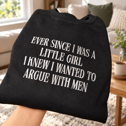 A folded black sweatshirt or sweatshirt shirt held up by a hand in a living room setting, featuring the text 'EVER SINCE I WAS A LITTLE GIRL I KNEW I WANTED TO ARGUE WITH MEN' printed in white serif letters.