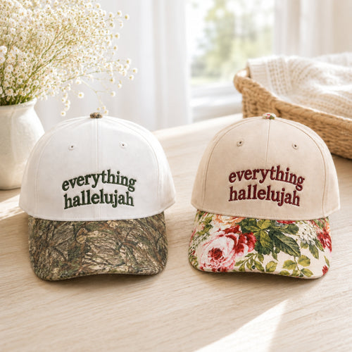 Two baseball caps on a light wood table in a bright indoor setting. The left cap is white on top with a camouflage-patterned brim and the embroidered text 'everything hallelujah' in dark green.