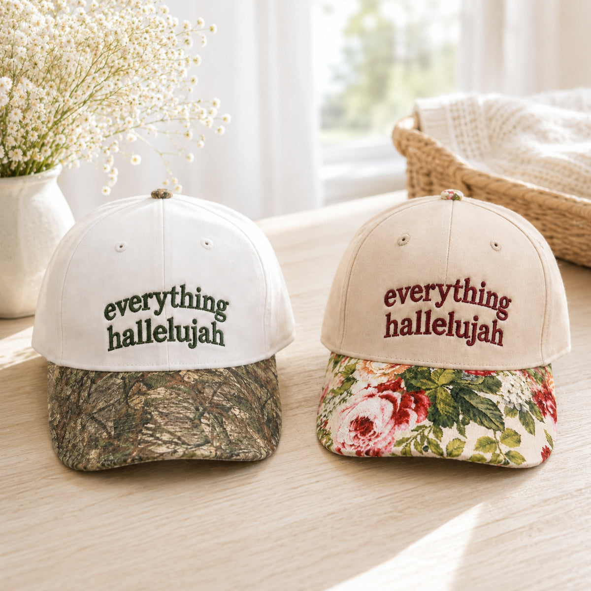 Two baseball caps on a light wood table in a bright indoor setting. The left cap is white on top with a camouflage-patterned brim and the embroidered text 'everything hallelujah' in dark green.