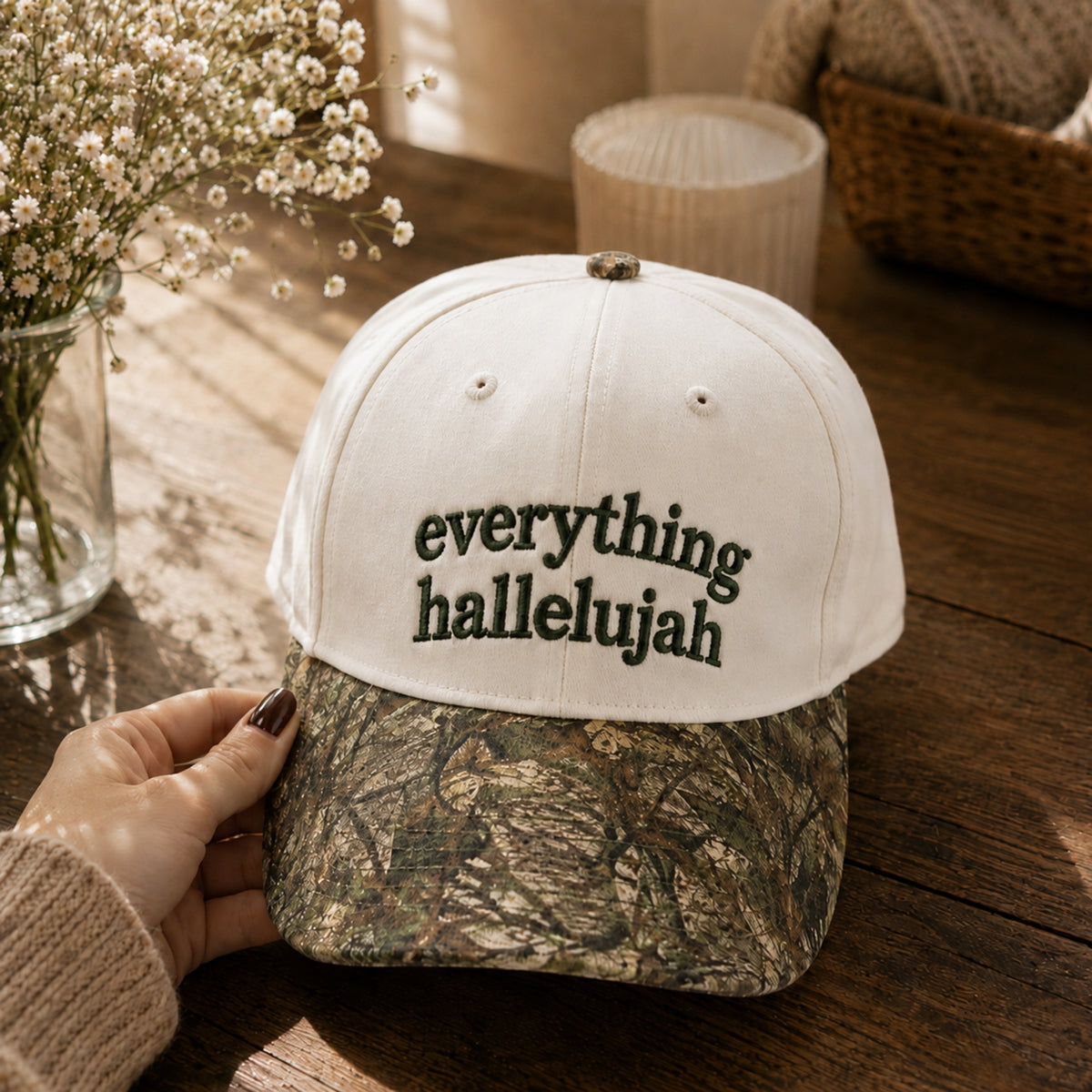 A white baseball cap with 'everything hallelujah' embroidered on the front, featuring a camouflage-patterned brim, held by a hand on a rustic wooden table with dried flowers in the background.