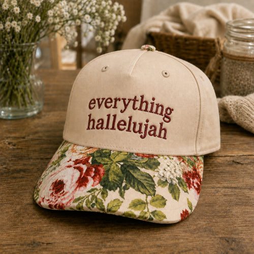 A beige baseball cap with 'everything hallelujah' embroidered in maroon on the front, featuring a floral-patterned brim with red, green, and cream flowers. The cap is styled on a rustic wooden table with a vase of baby's breath and a woven basket in the background.