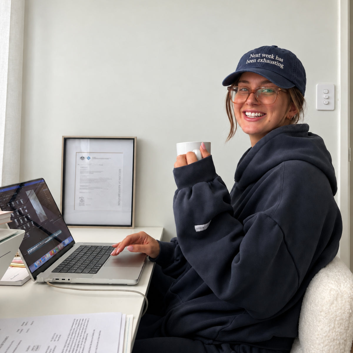A woman wearing a navy blue hoodie and a matching navy cap with 'Next Week, We Are Exciting' sits at a desk with a laptop and a mug, smiling in a bright indoor setting.