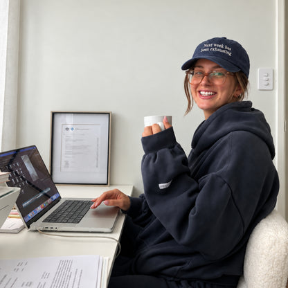 A woman wearing a navy blue hoodie and a matching navy cap with 'Next Week, We Are Exciting' sits at a desk with a laptop and a mug, smiling in a bright indoor setting.