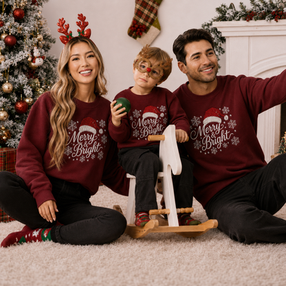 A family of three wearing matching maroon Christmas sweatshirts with 'Merry Bright' and snowflake prints, sitting together on a white rug in a decorated holiday living room.