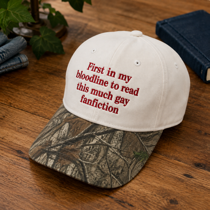 A baseball cap with a white crown and a camouflage-patterned brim, featuring red embroidered text that reads 'First in my bloodline to read this much gay fanfiction'. The cap is displayed on a wooden table with a book, jeans, and green leaves in the background.