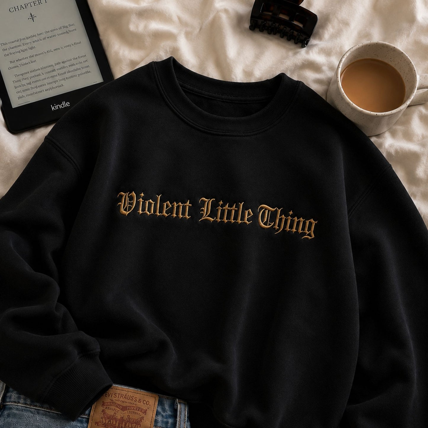 A black crewneck sweatshirt with 'Violent Little Thing' printed in a gold gothic font across the chest, styled in a cozy flat-lay arrangement with a Kindle, a cup of coffee, and jeans on a white textured blanket.