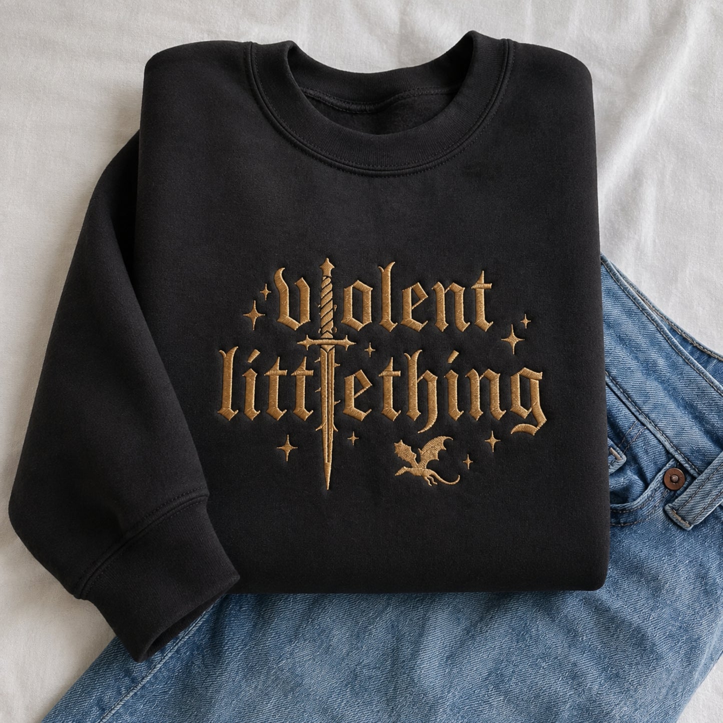 A folded black crewneck sweatshirt with a gold gothic-style graphic reading 'Violent LittleThing' with a sword, stars, and a small dragon, styled on top of blue jeans against a white fabric background.