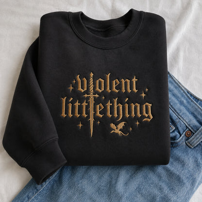A folded black crewneck sweatshirt with a gold gothic-style graphic reading 'Violent LittleThing' with a sword, stars, and a small dragon, styled on top of blue jeans against a white fabric background.