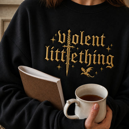 A person wearing a black sweatshirt with a gold gothic-style graphic reading 'Violent Littleshopping' across the chest, holding a white mug and a brown notebook in a cozy indoor setting.