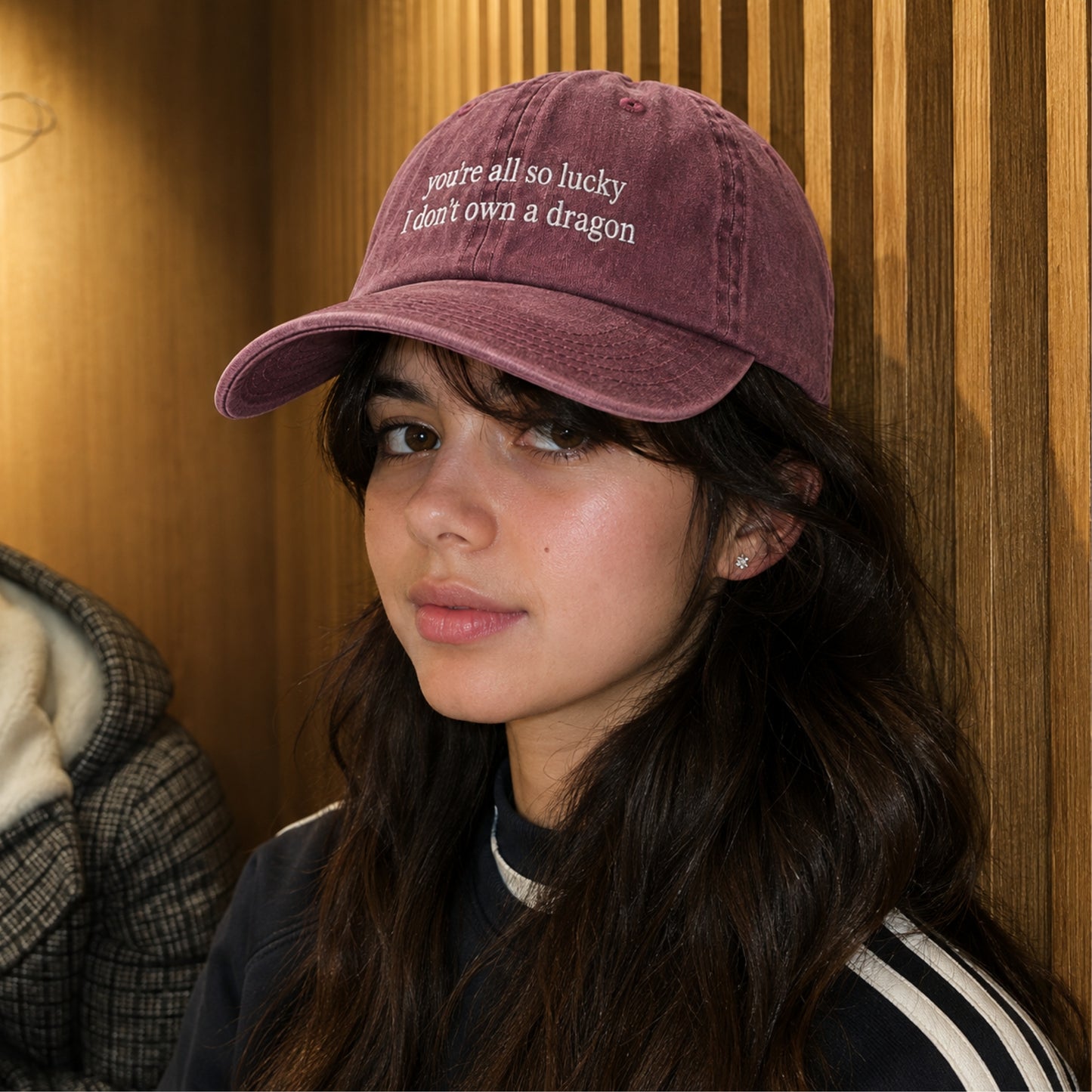 A woman wearing a dusty mauve baseball cap with white embroidered text that reads 'you're all so lucky I don't own a dragon'. She is posing in front of a warm wood-paneled wall.