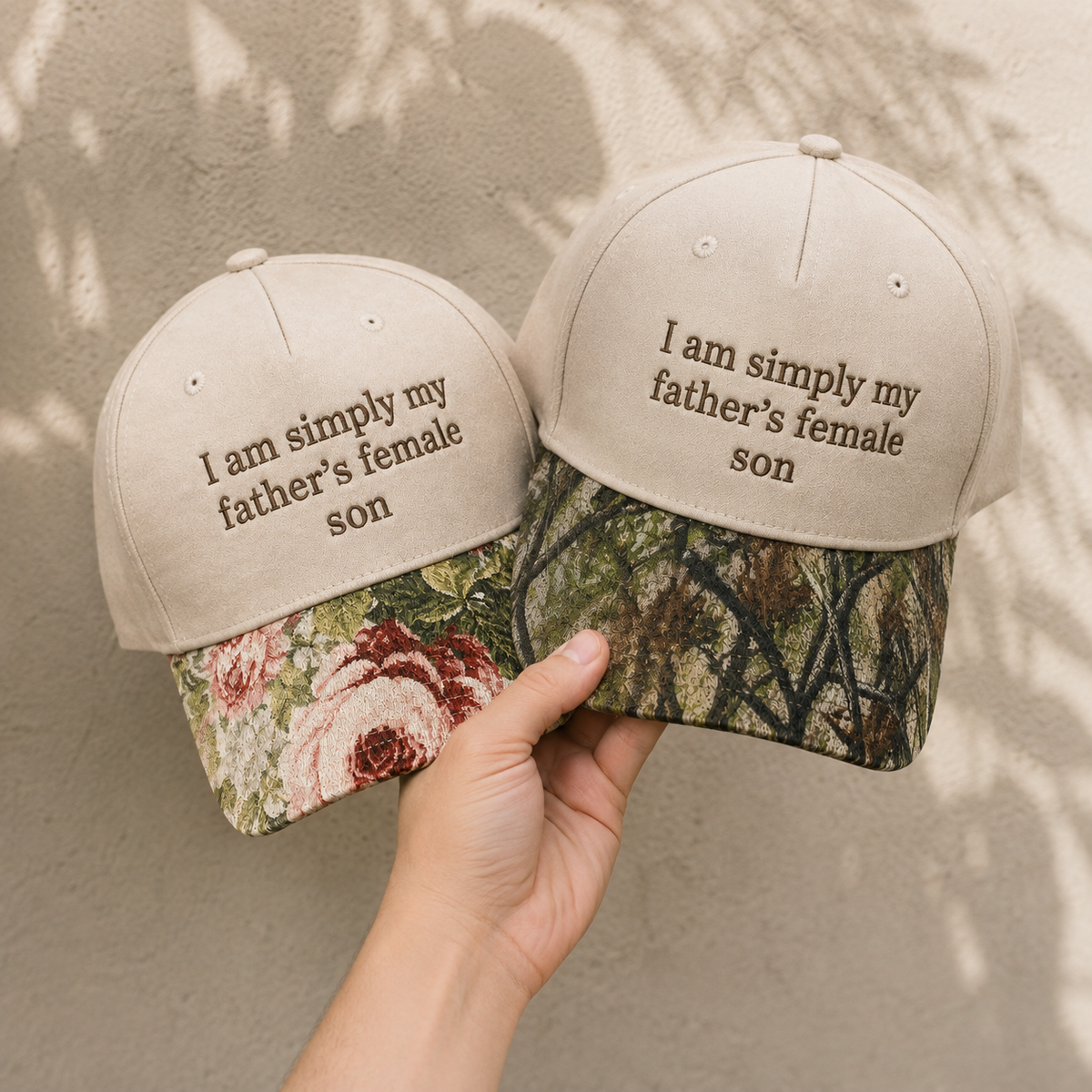 Two beige baseball caps with embroidered text reading 'I am simply my father's female son' are shown, one with a floral brim and one with a camouflage brim, held in a hand against a textured wall with leaf shadows.