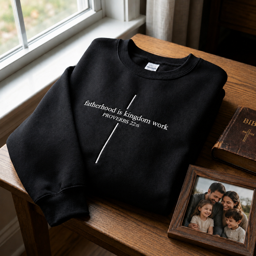 A black crewneck sweatshirt featuring a white cross graphic and text reading "fatherhood is kingdom work PROVERBS 22:6", styled on a wooden table next to a Bible and framed family photo.