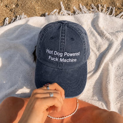 A navy blue washed baseball cap with white embroidered text reading 'Hot Dog Powered Fuck Machine' is worn by a person lying on a white fringed blanket outdoors on sand.