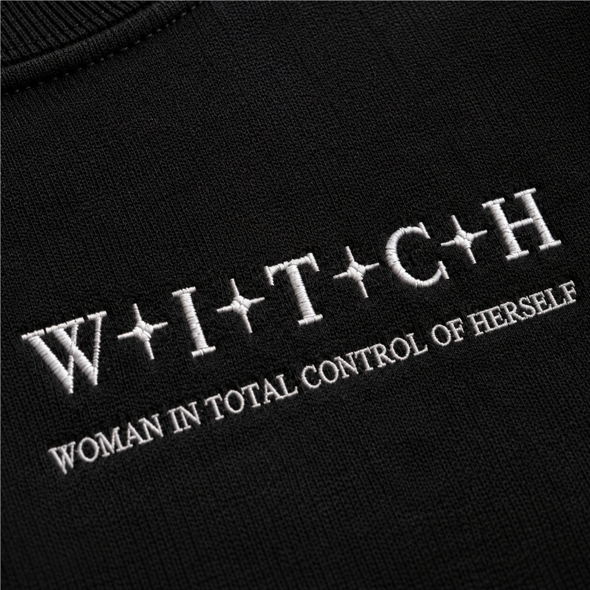 Close-up of a black knit garment with white embroidered text reading 'W+I+T+C+H' and 'WOMAN IN TOTAL CONTROL OF HERSELF'.