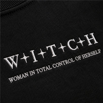 Close-up of a black knit garment with white embroidered text reading 'W+I+T+C+H' and 'WOMAN IN TOTAL CONTROL OF HERSELF'.