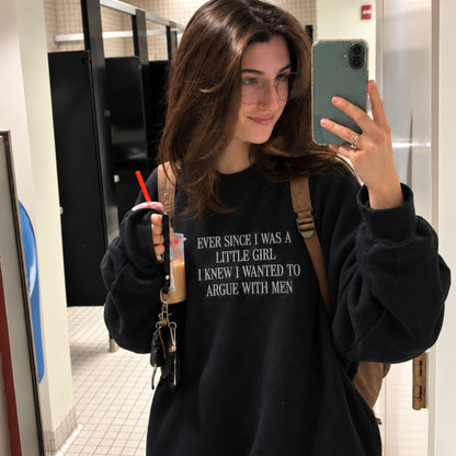 A woman taking a mirror selfie in a bathroom stall area, wearing a black oversized sweatshirt with white text that reads 'EVER SINCE I WAS A LITTLE GIRL I KNEW I WANTED TO ARGUE WITH MEN'. She is holding a drink and wearing a brown shoulder bag.