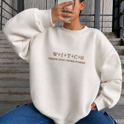 A person wearing an oversized cream sweatshirt with brown text reading 'W+I+T+C+H' and 'WOMAN IN TOTAL CONTROL OF HERSELF' on the chest, paired with blue jeans, seated on outdoor concrete steps.