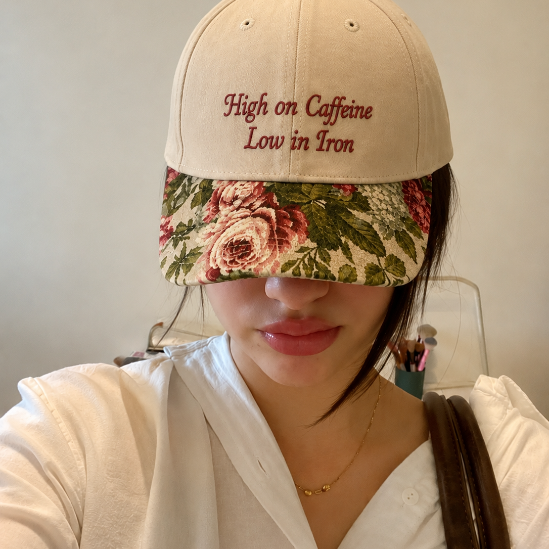 Close-up selfie of a woman wearing a beige baseball cap with red embroidered text 'High on Caffeine Low in Iron' and a vintage floral rose tapestry visor.