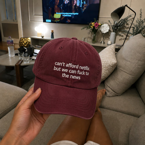 A person holds a maroon baseball cap with white embroidered text reading 'can't afford netflix but we can fuck to the news' in a living room setting.