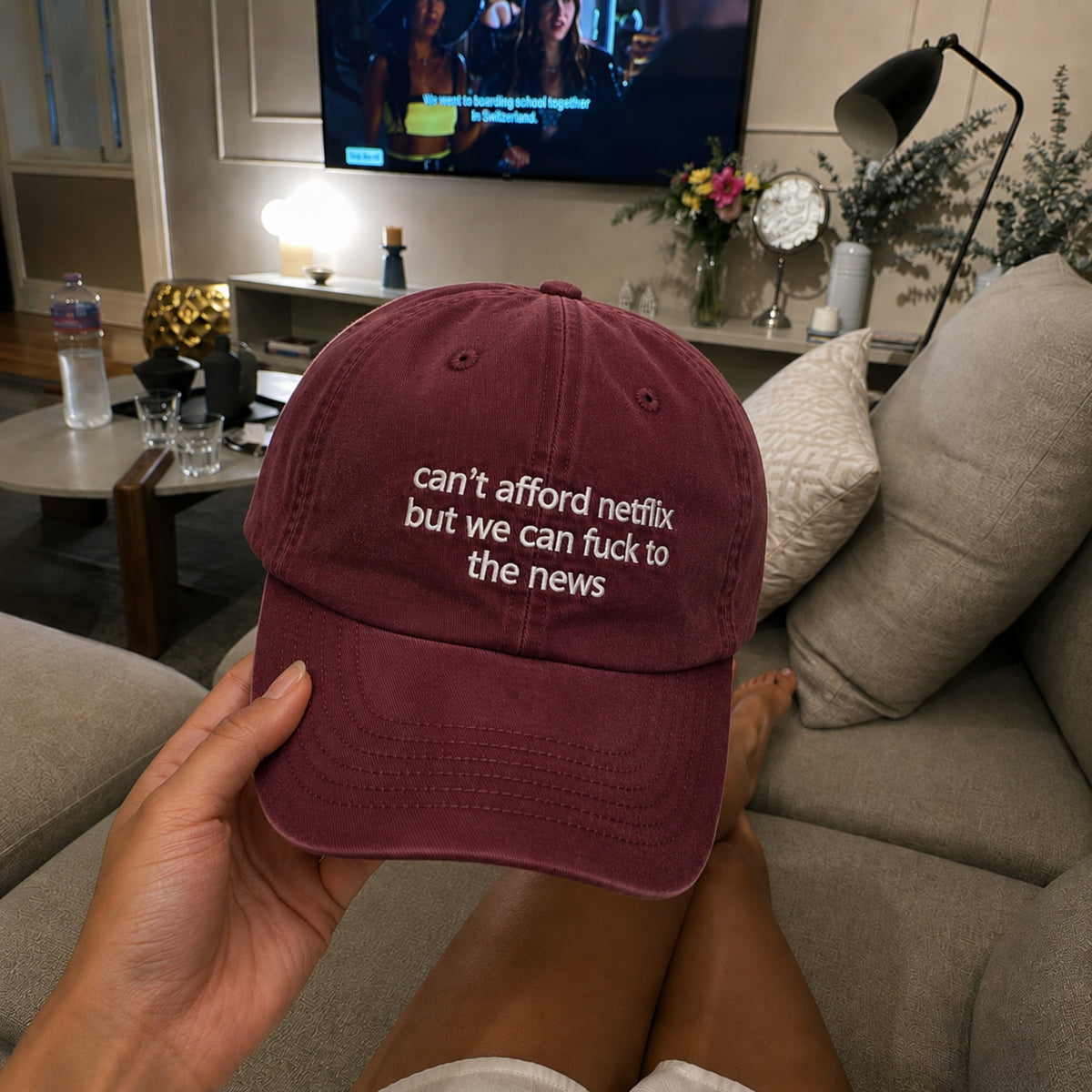 A person holds a maroon baseball cap with white embroidered text reading 'can't afford netflix but we can fuck to the news' in a living room setting.