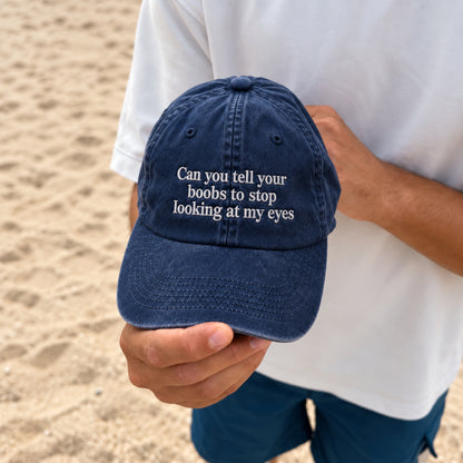 A navy blue baseball cap with white embroidered text reading 'Can you tell your boobs to stop looking at my eyes', held by a person wearing a white t-shirt and blue shorts on a sandy beach.