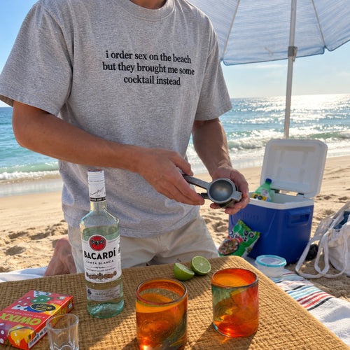 A man on a beach wearing a light gray graphic t-shirt with a humorous print, holding a lime squeezer over a table with a bottle of Bacardi rum and colorful drinks. A blue cooler and beach umbrella are in the background.