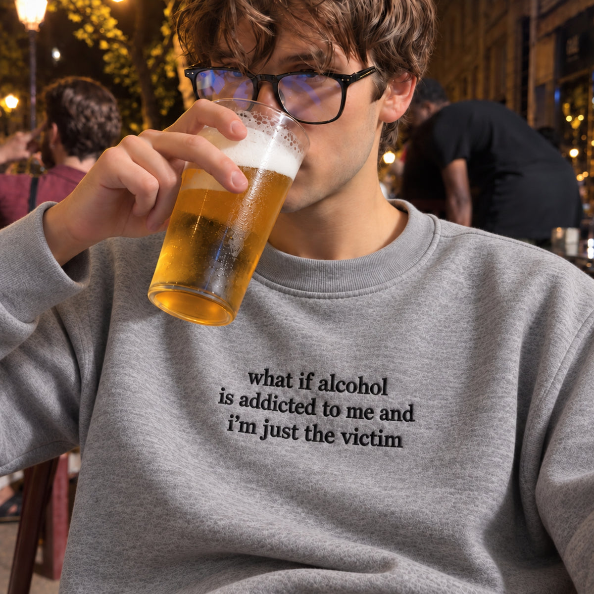 A man wearing black-rimmed glasses and a gray crewneck sweatshirt with the text 'what if alcohol is addicted to me and i'm just the victim' drinking a pint of beer in a dimly lit bar setting.