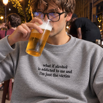 A man wearing black-rimmed glasses and a gray crewneck sweatshirt with the text 'what if alcohol is addicted to me and i'm just the victim' drinking a pint of beer in a dimly lit bar setting.