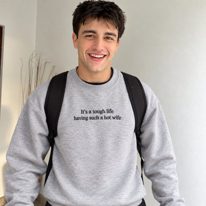 A smiling young man wearing a light gray crewneck sweatshirt with black text that reads 'It's a tough life having such a hot wife'. He has a black backpack on and is standing against a plain off-white wall.