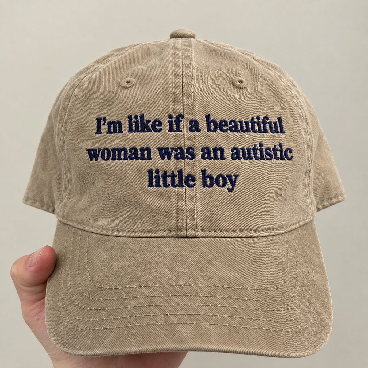 A beige cotton baseball cap held by a hand, featuring navy blue embroidered text that reads 'I'm like if a beautiful woman was an autistic little boy'.