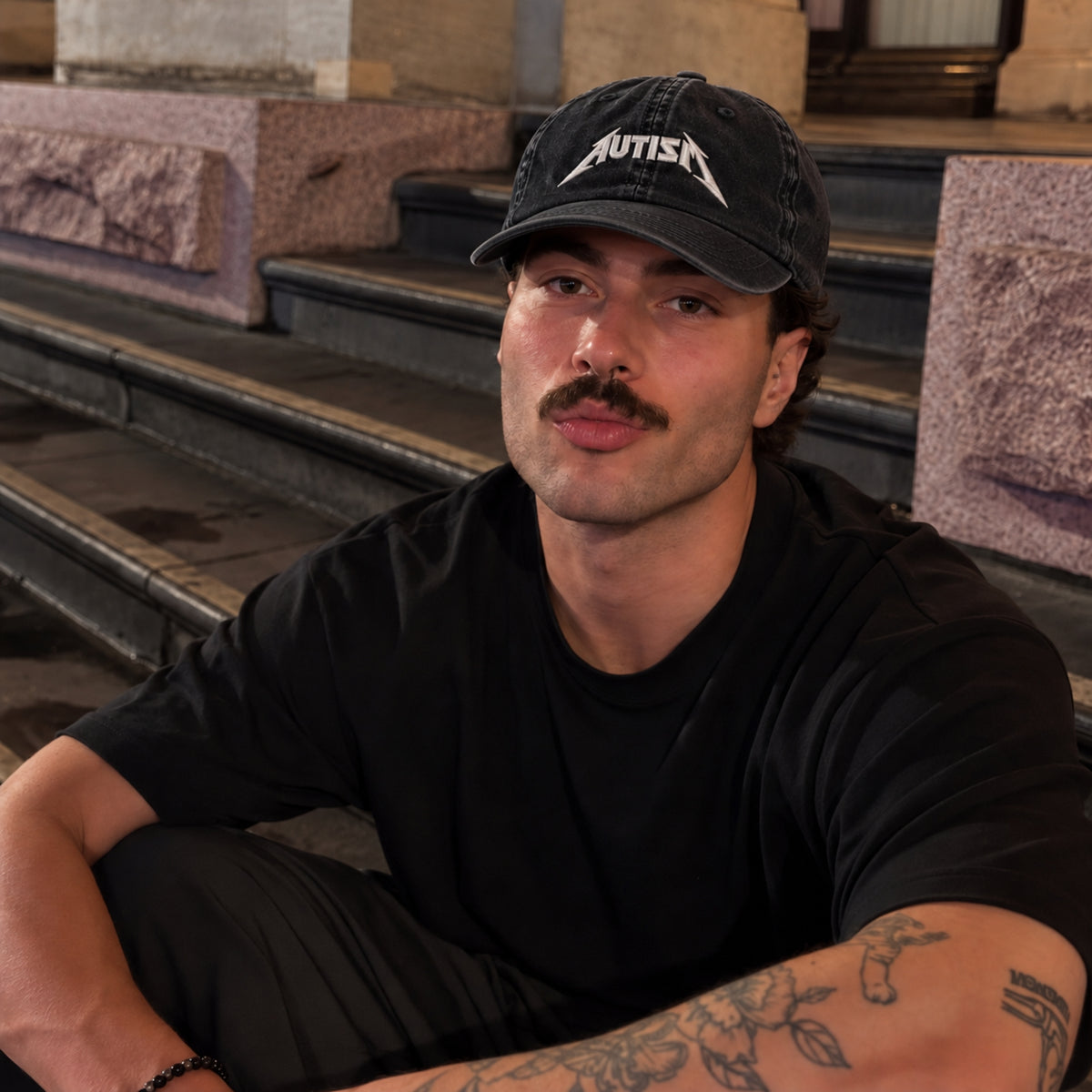 A man wearing a black t-shirt and a black baseball cap with the word 'AUTISM' in white text, sitting on stone steps.
