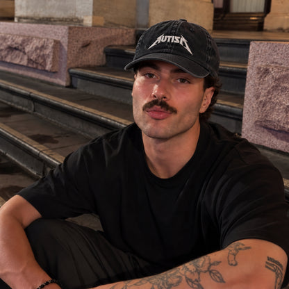 A man wearing a black t-shirt and a black baseball cap with the word 'AUTISM' in white text, sitting on stone steps.