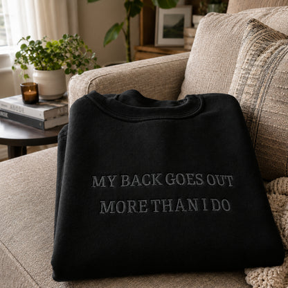 A black crewneck sweatshirt with the text 'MY BACK GOES OUT MORE THAN I DO' in gray embroidered lettering, folded and displayed on a beige fabric couch in a cozy living room setting.
