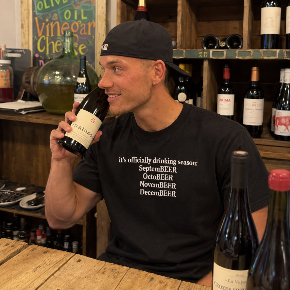 A man wearing a black t-shirt with white text reading 'it's officially drinking season: SeptemBEER OctoBEER NovemBEER DecemBEER' and a black baseball cap, holding a wine bottle in a wine shop setting.