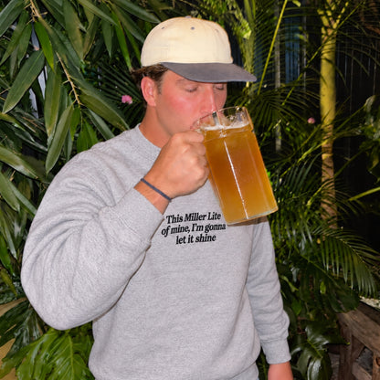 Male model wearing a sport grey sweatshirt with black embroidered text reading “This Miller Lite of mine, I’m gonna let it shine,” drinking from a large beer mug in a tropical outdoor setting.