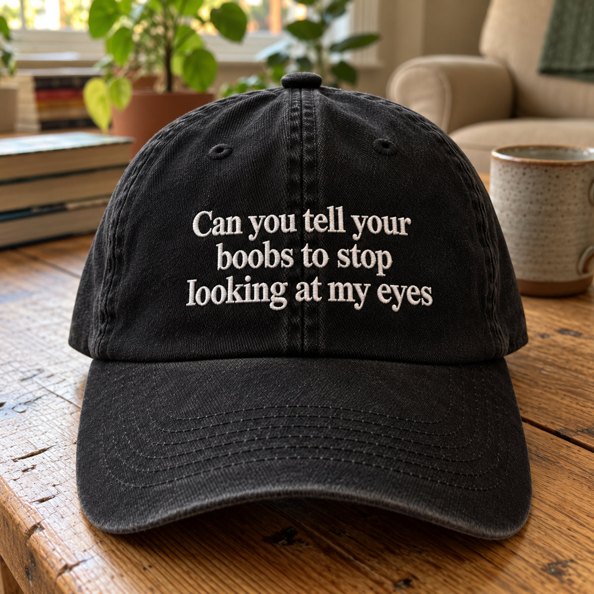 A black washed cotton baseball cap with white embroidered text reading 'Can you tell your boobs to stop looking at my eyes' sits on a rustic wooden table in a cozy indoor setting with a mug and plants in the background.