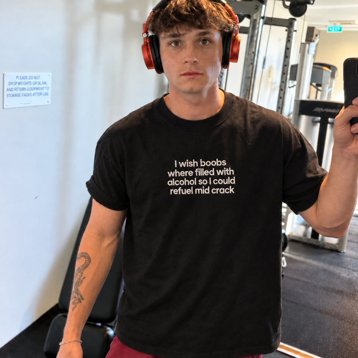 A man in a gym wearing a black graphic t-shirt with white text that reads 'I wish boobs where filled with alcohol so I could refuse mid crack'. He is also wearing orange headphones and holding a phone.