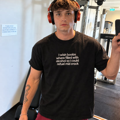 A man in a gym wearing a black graphic t-shirt with white text that reads 'I wish boobs where filled with alcohol so I could refuse mid crack'. He is also wearing orange headphones and holding a phone.