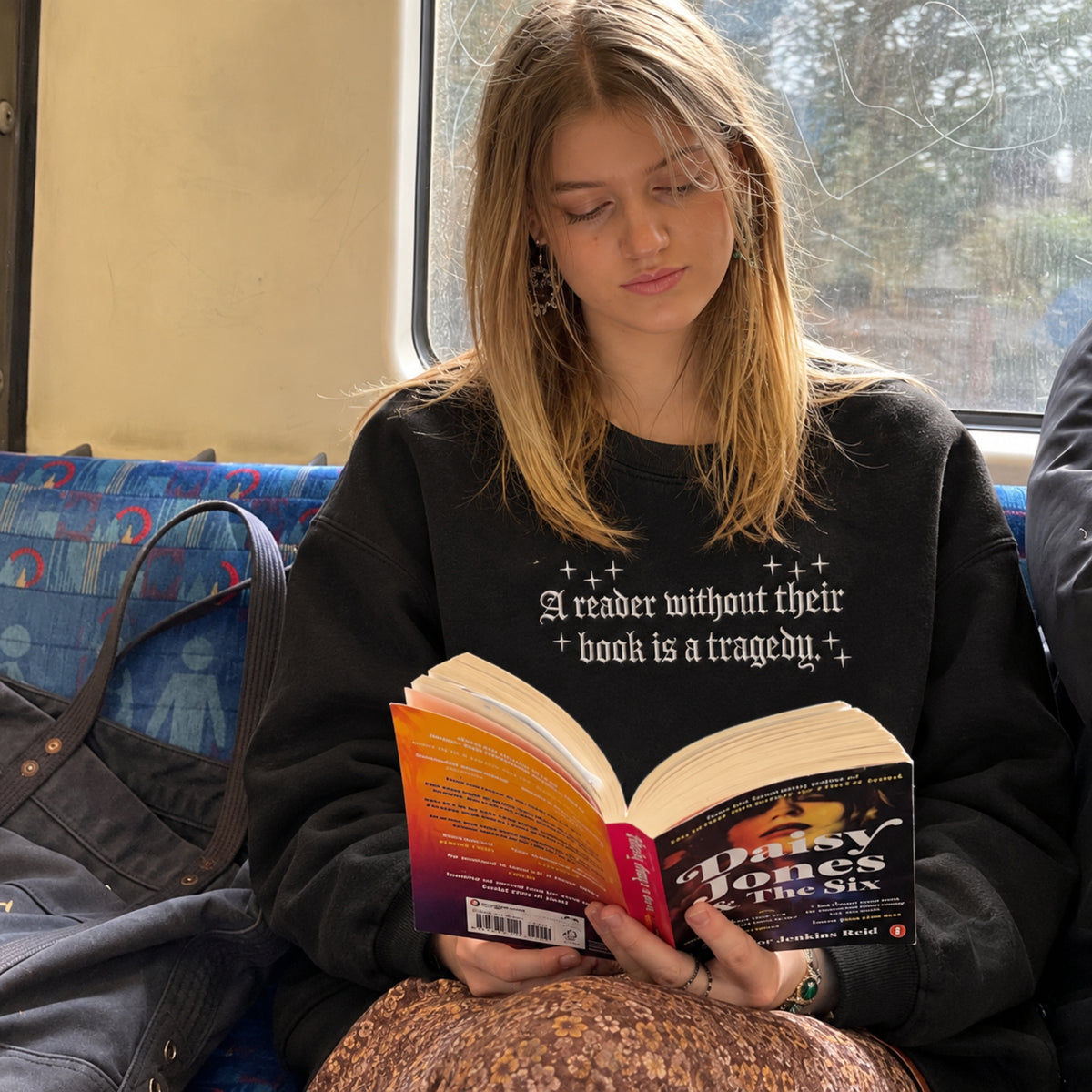A young woman with blonde hair sits on a train, reading a book. She is wearing a black crewneck sweatshirt with the white printed text 'A reader without their book is a tragedy.