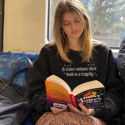 A young woman with blonde hair sits on a train, reading a book. She is wearing a black crewneck sweatshirt with the white printed text 'A reader without their book is a tragedy.