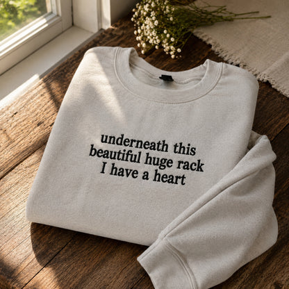 A folded off-white crewneck sweatshirt with black text reading 'underneath this beautiful huge rack I have a heart', styled on a wooden windowsill with a small bouquet of baby's breath flowers nearby.