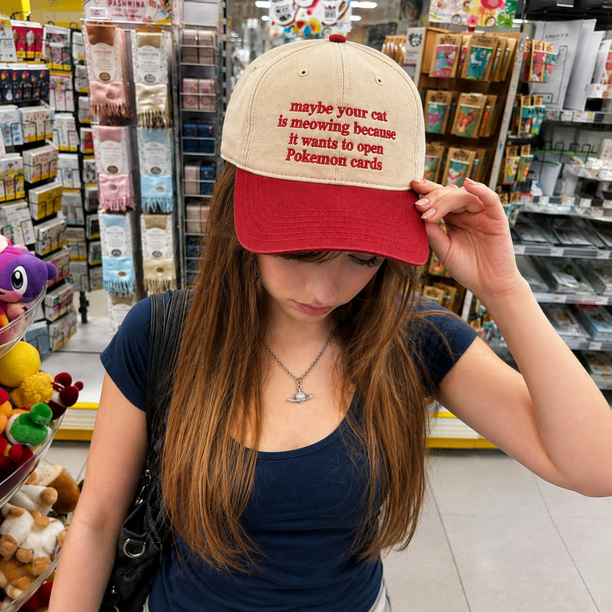 A woman in a retail store wearing a beige and red baseball cap with red embroidered text that reads 'maybe your cat is meowing because it wants to open Pokemon cards'.