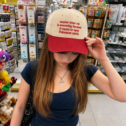 A woman in a retail store wearing a beige and red baseball cap with red embroidered text that reads 'maybe your cat is meowing because it wants to open Pokemon cards'.