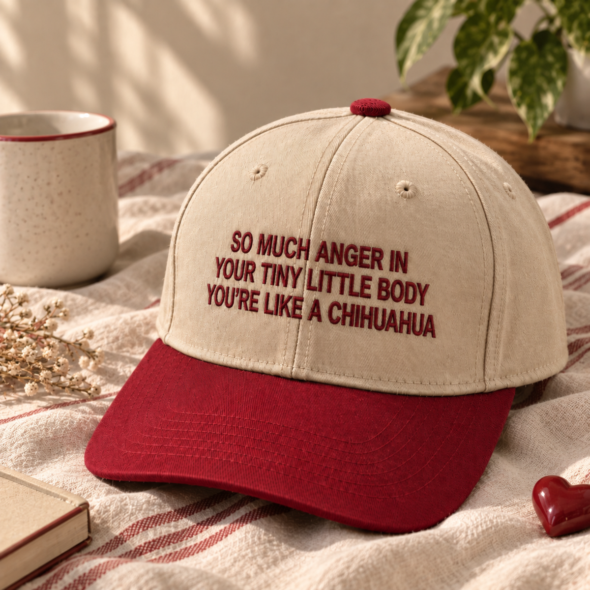 A baseball cap with a cream-colored crown and a red brim, featuring the embroidered text 'SO MUCH ANGER IN YOUR TINY LITTLE BODY YOU'RE LIKE A CHIHUAHUA' in red. The cap is styled on a red and white striped blanket with a mug, dried flowers, and a book in the background.