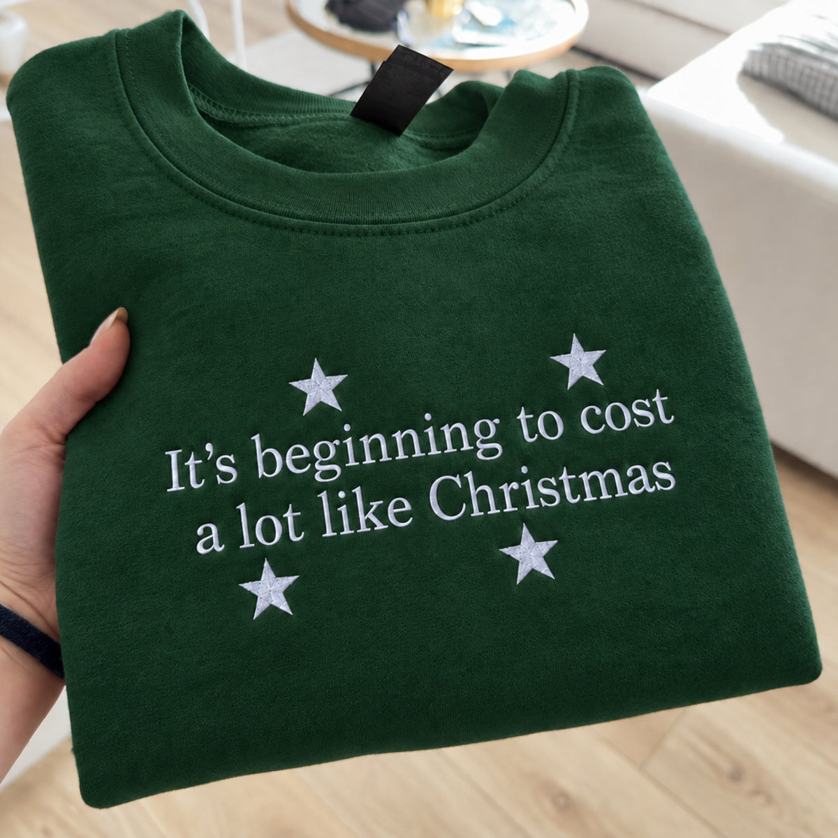 A dark green crewneck sweatshirt folded and held by a hand, featuring the white embroidered text 'It's beginning to cost a lot like Christmas' surrounded by white stars.