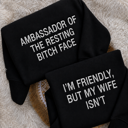 Two folded black sweatshirts with white embroidered text, displayed on a textured beige faux fur surface. The top sweatshirt reads 'AMBASSADOR OF THE RESTING BITCH FACE' and the bottom sweatshirt reads 'I'M FRIENDLY, BUT MY WIFE ISN'T'.