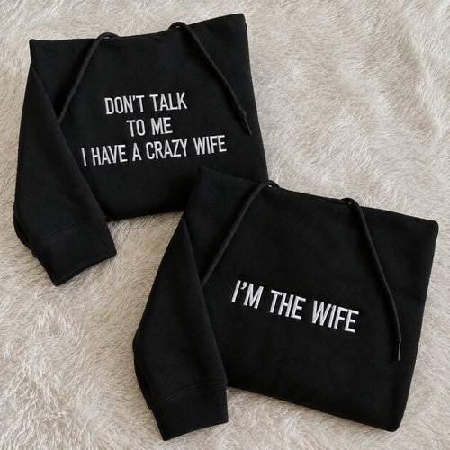 Two black hoodies laid flat on a beige faux-fur surface. One hoodie reads 'DON'T TALK TO ME I HAVE A CRAZY WIFE' and the other reads 'I'M THE WIFE' in white text.