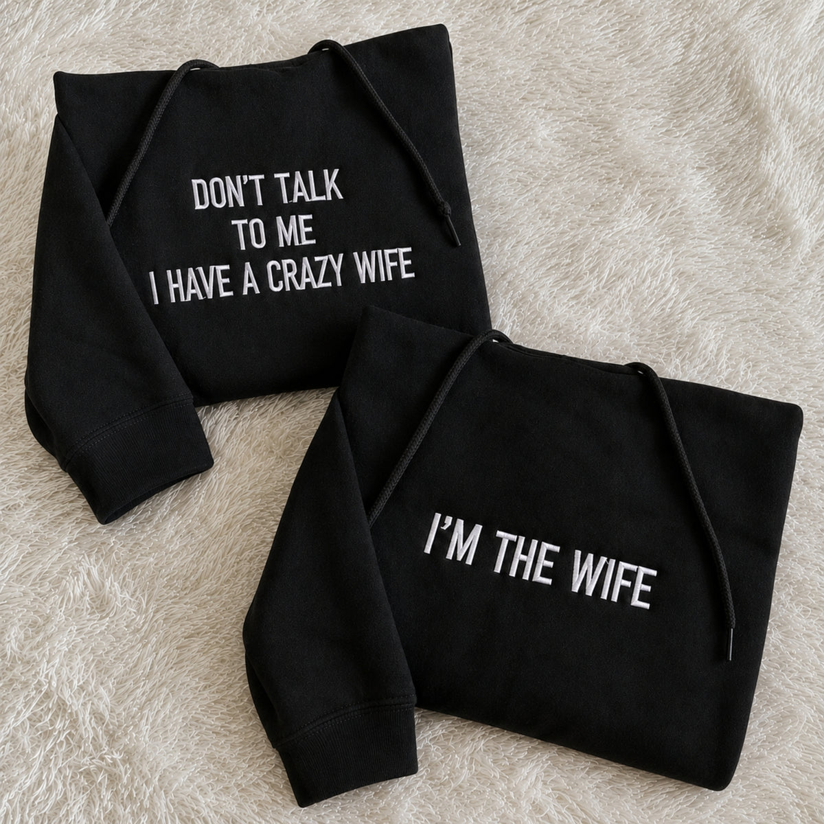 Two black hoodies laid flat on a beige faux-fur surface. One hoodie reads 'DON'T TALK TO ME I HAVE A CRAZY WIFE' and the other reads 'I'M THE WIFE' in white text.
