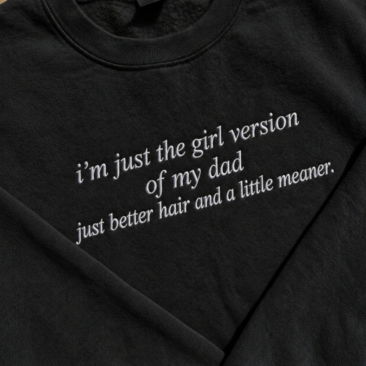 Close-up of a black crewneck t-shirt featuring white embroidered text that reads 'i'm just the girl version of my dad just better hair and a little meaner.'.